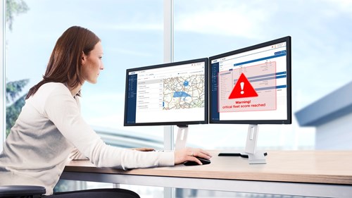A fleet manager sits at her workstation in front of two screens. The left screen shows a map and the right screen displays a warning in the VDO Tachograph Management software.