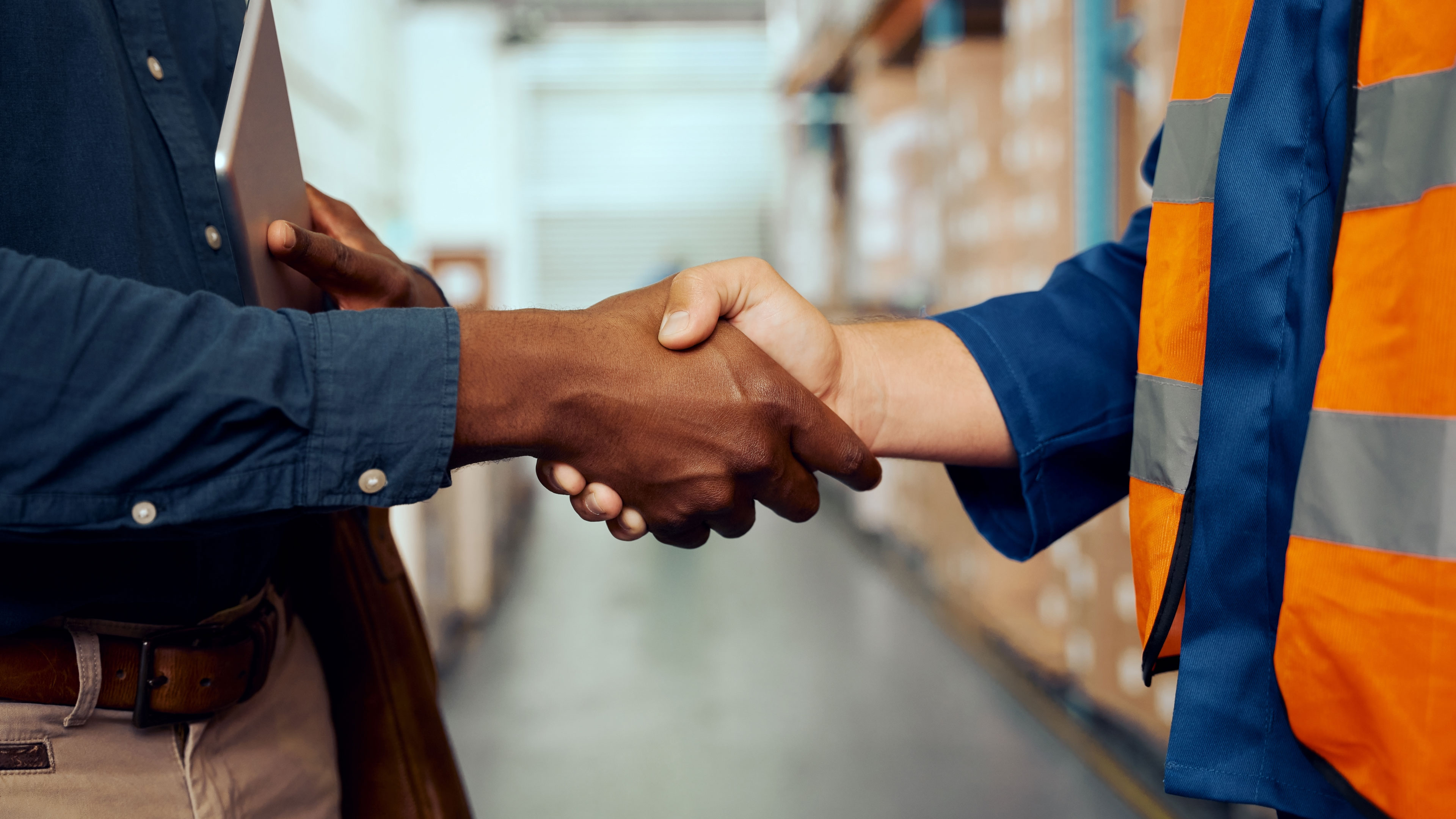 Closeup of two men shaking hands. One is causasian and is wearing a high-visibility vest. The other one is black, dressed in khakis and holding a tablet computer in his other hand.